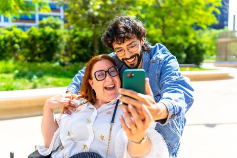 Cheerful couple taking a selfie in a park, the woman sitting on a wheelchair and the man embracing