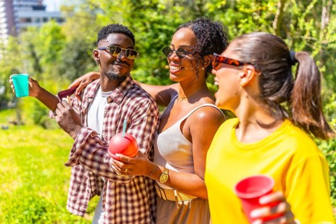 Three friends are walking through a park, enjoying drinks and each other's company on a sunny day