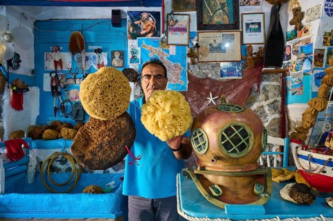 A man holds sponges next to an antique diving helmet in a blue, maritime-decorated room, Sponge