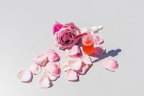 From above, a bottle of rose water serum with a dropper sits among delicate pink rose petals and a