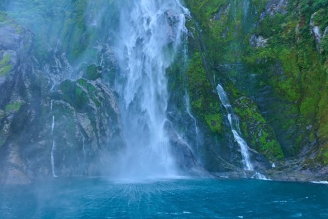 Waterfall pours over overgrown cliffs into the blue water, summer, Milford Sound, Te Anau,