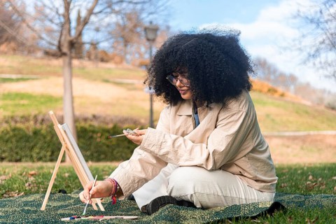 A young black woman with curly hair focuses on painting in a serene park, surrounded by vibrant