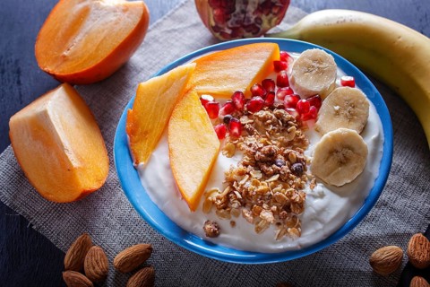 A blue plate with greek yogurt, granola, persimmon, banana, pomegranate black wooden background.