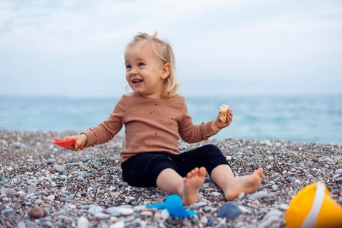A young child sits on a pebble beach, holding a snack in one hand. The child girl appears curious