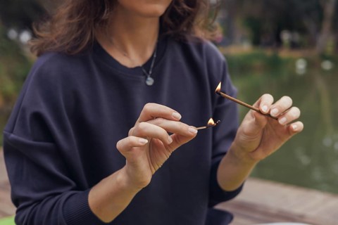 A young Caucasian woman with long brown hair sits by a pond, holding an incense stick. Smoke rises