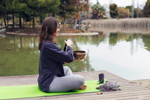 A young Caucasian woman with long brown hair sits cross-legged on a green yoga mat by a lake. She
