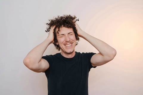 A young man with curly hair smiles widely, placing his hands on his head. He wears a casual black