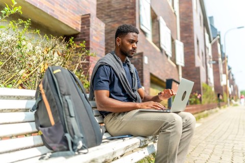 Side view portrait of a young african man working with laptop sitting on a city bench