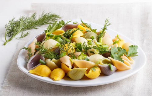 Conchiglie colored pasta with fresh greengrocery on a linen tablecloth on white wooden background.