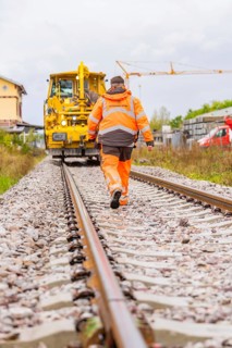 Workers in orange safety clothing walk along the tracks towards a yellow locomotive, tamping