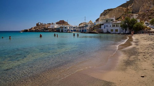 People bathe in clear water on a sandy beach with white buildings and a rocky bay, Palio Ergostasio