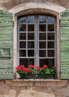 Old window with weathered green wooden shutters and red geraniums on a rustic wall, Provence,