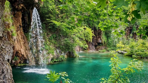 An enchanting waterfall pours into a turquoise river surrounded by lush foliage and rocks, Plitvice