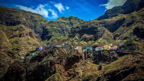Panoramic view, small colorful settlement nestled in raw, dramatic mountains under blue skies,