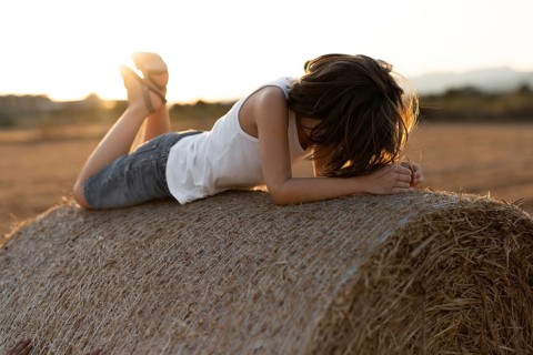 A child in a white tank top and denim shorts lies on a hay bale, enjoying a serene sunset in the