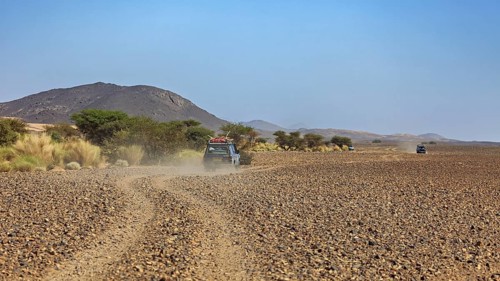 An off-road vehicle drives on a dusty road through a stony desert landscape with mountains in the