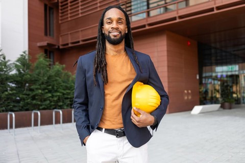 A cuban businessman stands confidently outside a modern building, holding a yellow hard hat. He is