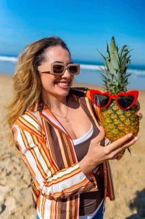 Woman wearing sunglasses and a striped shirt smiling while holding a fresh pineapple adorned with