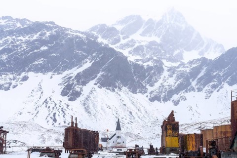 Norwegian wooden church between rusted industrial plants of the former whaling station, snow-capped