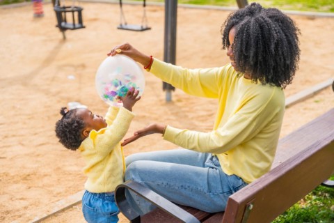 African mother and daughter playing with a balloon sitting on a park bench outdoors