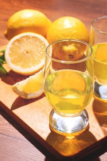 Two clear glasses sit on a wooden board filled with yellow lemon drink. Whole and sliced lemons are