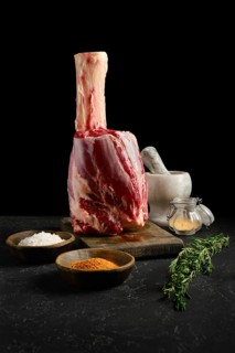 A large cut of beef hindshank on a wooden board with salt, spices, and herbs nearby. A mortar and