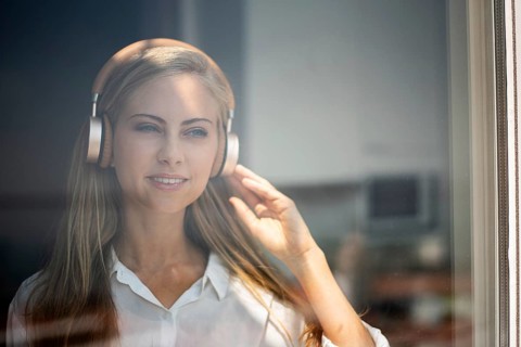 A woman stands by a window, wearing headphones and enjoying music. She looks relaxed and focused.