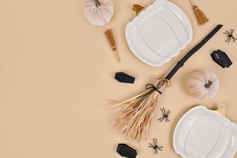 Halloween party flat lay with pumpkin shaped plates, witch broom, spiders and coffins on beige