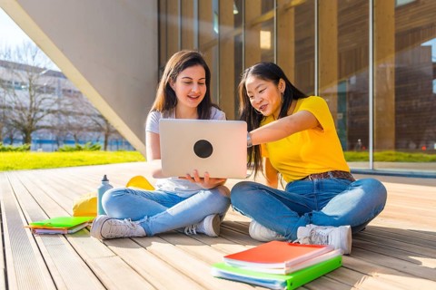 Asian and caucasian students using laptop in the campus sitting next to the university building