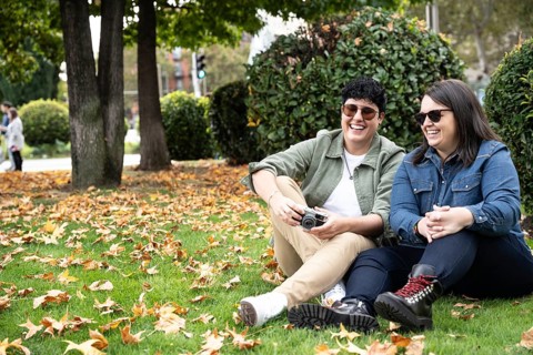 A joyful lesbian couple sits amidst autumn leaves in a park, sharing laughter. With casual attire