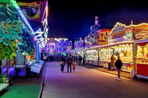 An illuminated fairground street at night with people walking along colorful stalls, carousel and