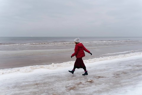A woman in red walks in winter along the seashore, A person in a red coat walks alone along a snowy
