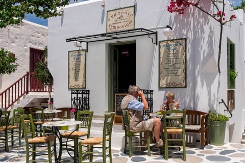 Two people sitting in a cosy outdoor café in sunny weather, surrounded by white buildings and