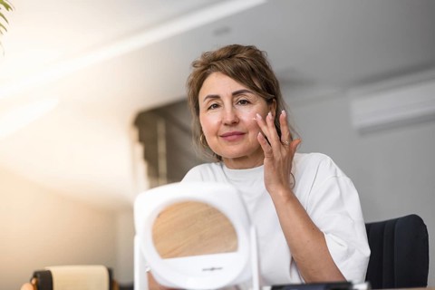 A woman is carefully applying moisturizer to her face while sitting at a table in a relaxed indoor