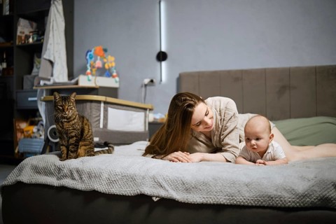 A mother lies on her stomach on a bed next to her infant, who is looking around. A cat sits nearby.