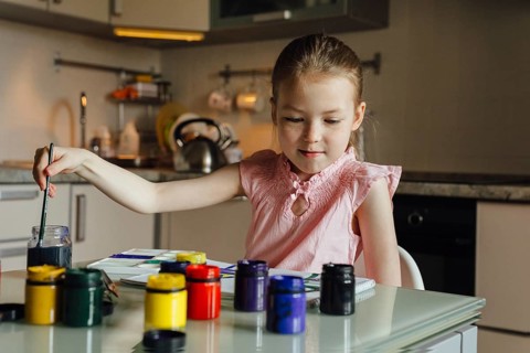 A young girl in a pink top paints creatively in a modern kitchen. Surrounded by colorful paint