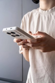 A teenager stands indoors using a smartphone with a white protective case, possibly typing or
