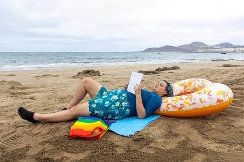 A transgender man lies on the sandy shore whilst engaging in a book, casually propped against a