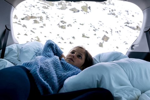 A girl lies comfortably in the back of a car surrounded by pillows and blankets. Outside, a snowy