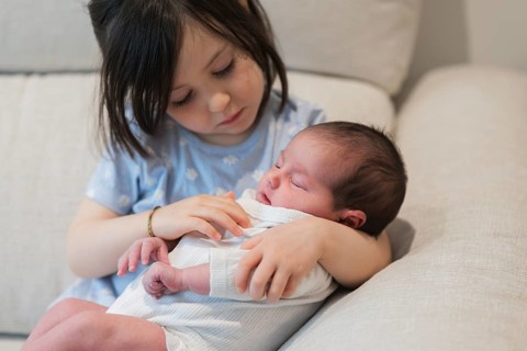 A young girl gently cradles her newborn sibling on a comfy sofa, highlighting the special bond they