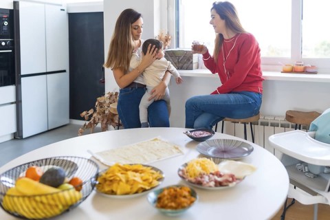 Two mothers are chatting and drinking juice in a modern kitchen, preparing a meal of tacos and