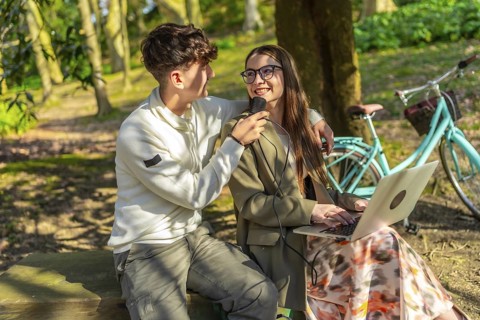 Two young journalists are recording a podcast in a park, using a microphone and laptop, with a