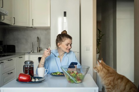 A woman sits at a kitchen table, enjoying a fresh salad while checking her phone. A curious cat