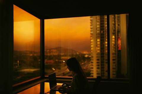 Side view of an unrecognizable woman sitting at a desk, working on a laptop in a hotel room with a
