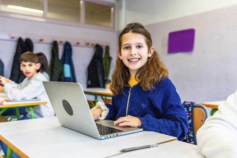 Elementary school student smiling and using laptop during class, with classmates and coats hanging