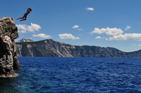 Person jumps off a cliff into the ice-cold water of Crater Lake, the mountain backdrop is visible,