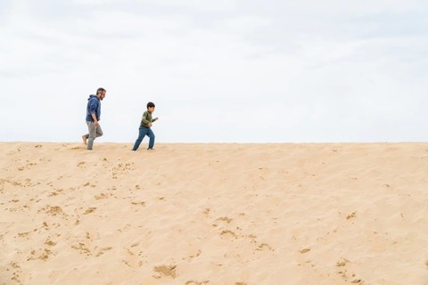 A father and son enjoy a carefree walk on a vast, sandy beach under a cloudy sky. The image