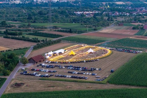 Aerial view of an illuminated festival in a field surrounded by cars and surrounding landscape,