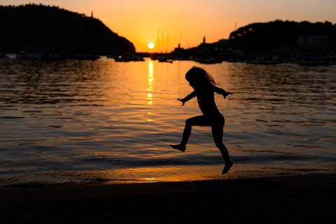 A child is joyfully jumping near the water as the sun sets, casting a beautiful silhouette against