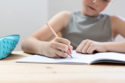 A young student concentrates on writing in a notebook with a pencil, a pencil case resting on the
